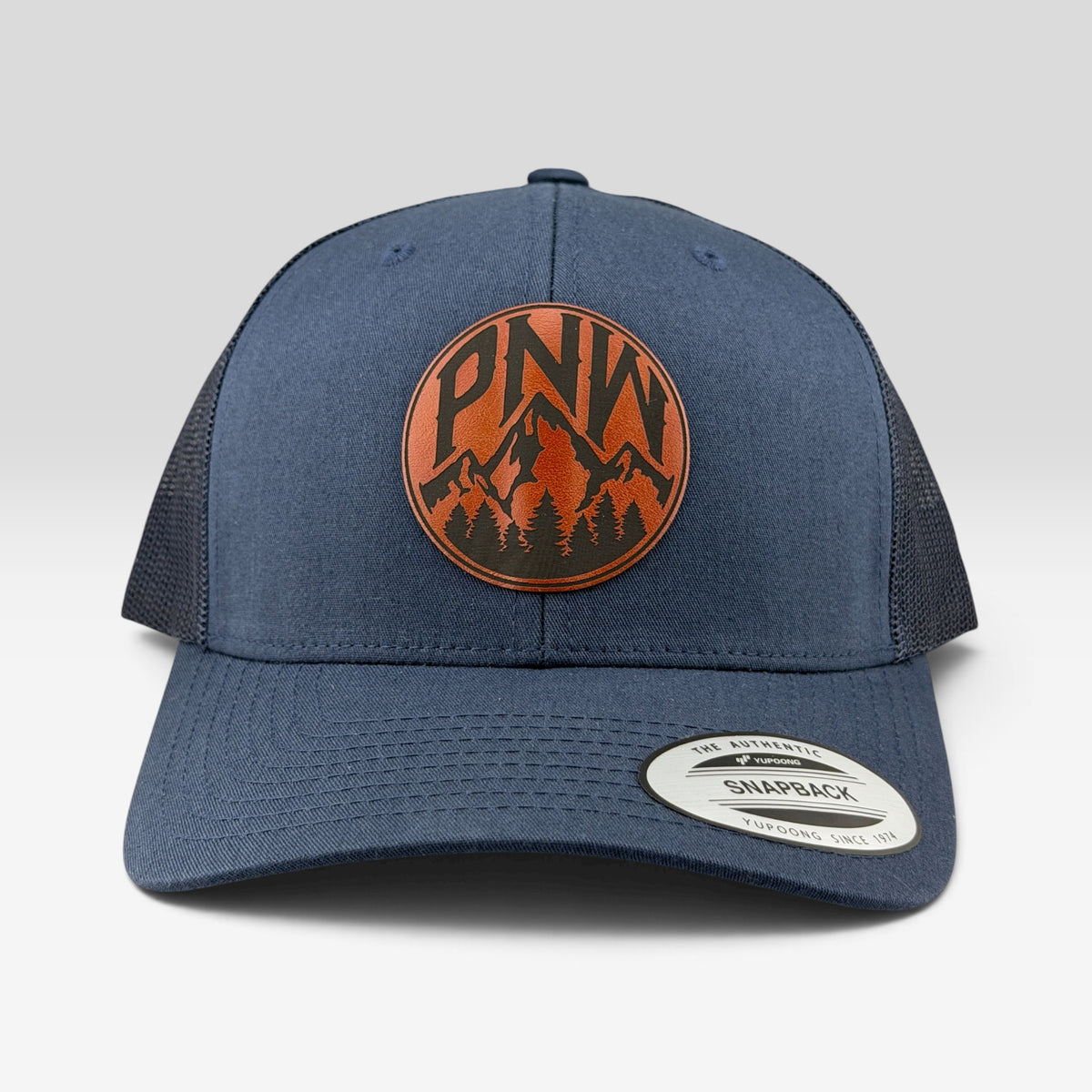 A navy blue trucker hat with a circular orange and black mountain logo patch on the front that reads 'PNW'. The brim has a visible Snapback sticker, and the background is a clean light gray studio backdrop.