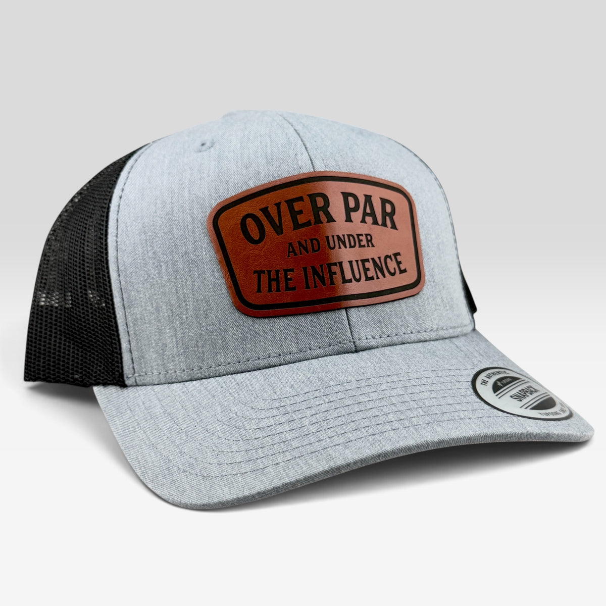 A light heather gray trucker hat with black mesh back panels and a brown leather patch on the front that reads 'OVER PAR AND UNDER THE INFLUENCE'.