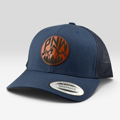 A navy blue trucker-style snapback cap with a circular brown and black mountain logo patch on the front. The cap features a structured navy crown, a matching navy curved brim, and dark mesh back panels.