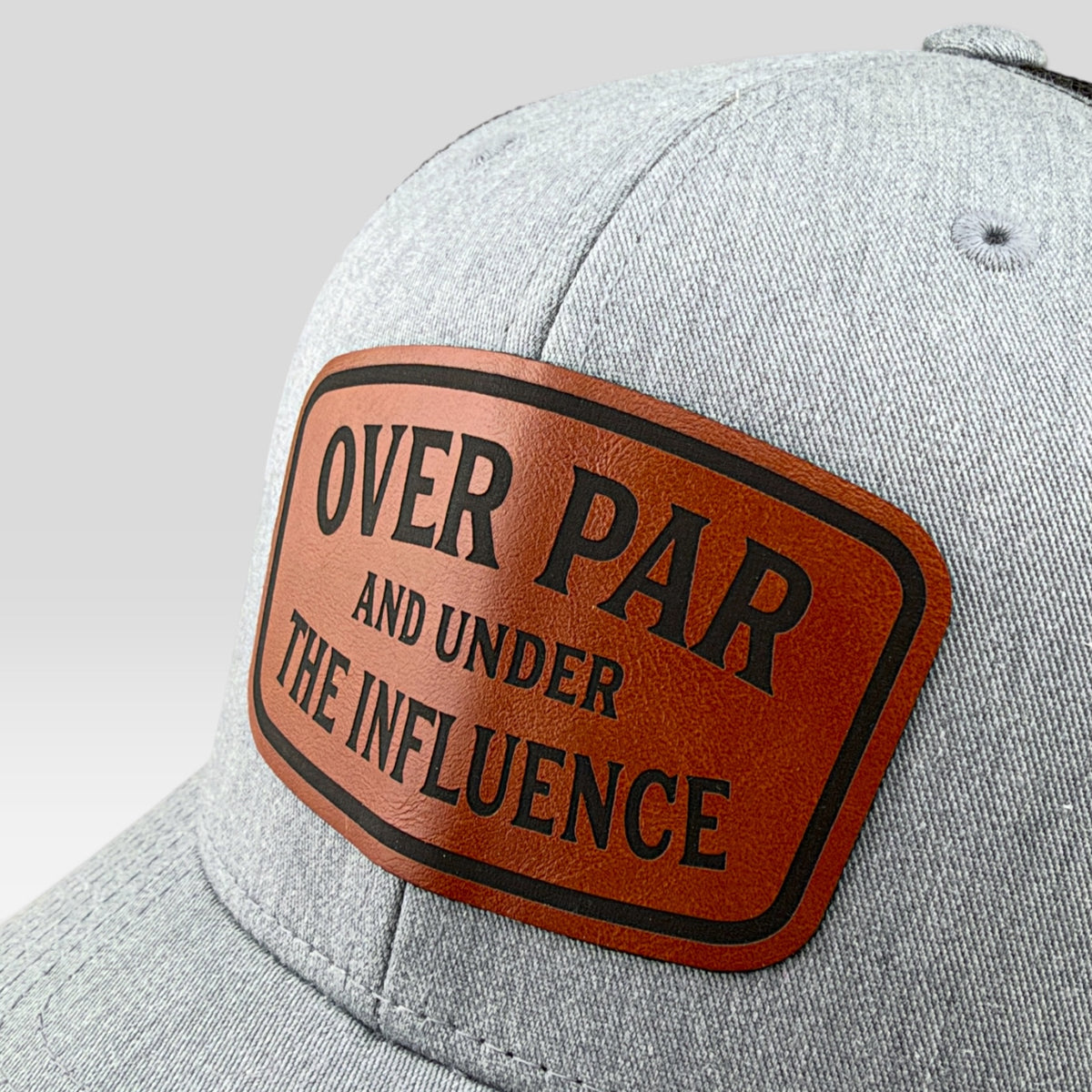 A close-up of a heather gray baseball cap featuring a brown leather patch with black text that reads 'OVER PAR AND UNDER THE INFLUENCE'.