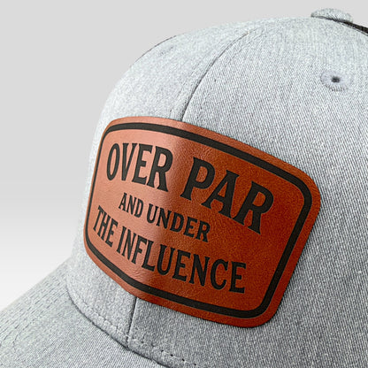 A close-up of a heather gray baseball cap featuring a brown leather patch with black text that reads 'OVER PAR AND UNDER THE INFLUENCE'.