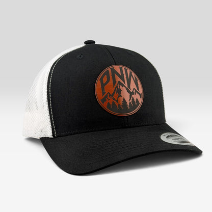 A black and white trucker hat featuring a circular brown leather patch on the front with the letters 'PNW' and a mountain and pine tree graphic. The brim has a silver metal sticker.
