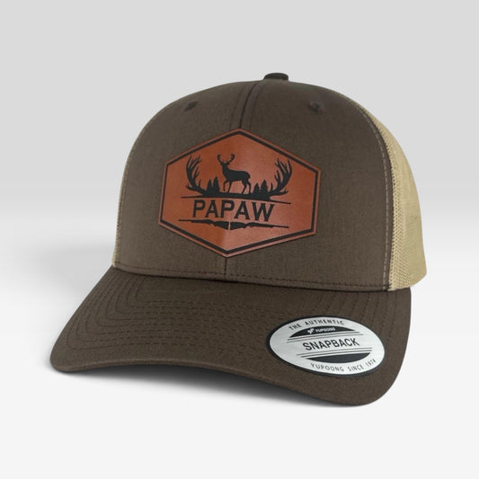 A brown and beige trucker hat with a brown leather front patch featuring a deer silhouette and the word 'PAPAW'.