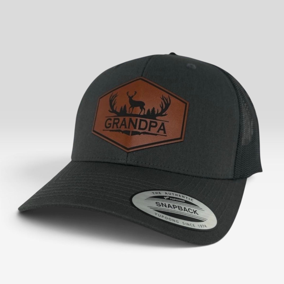 A dark charcoal gray trucker hat with a brown leather patch on the front featuring a deer and the word 'GRANDPA'. The brim has a white Snapback sticker.