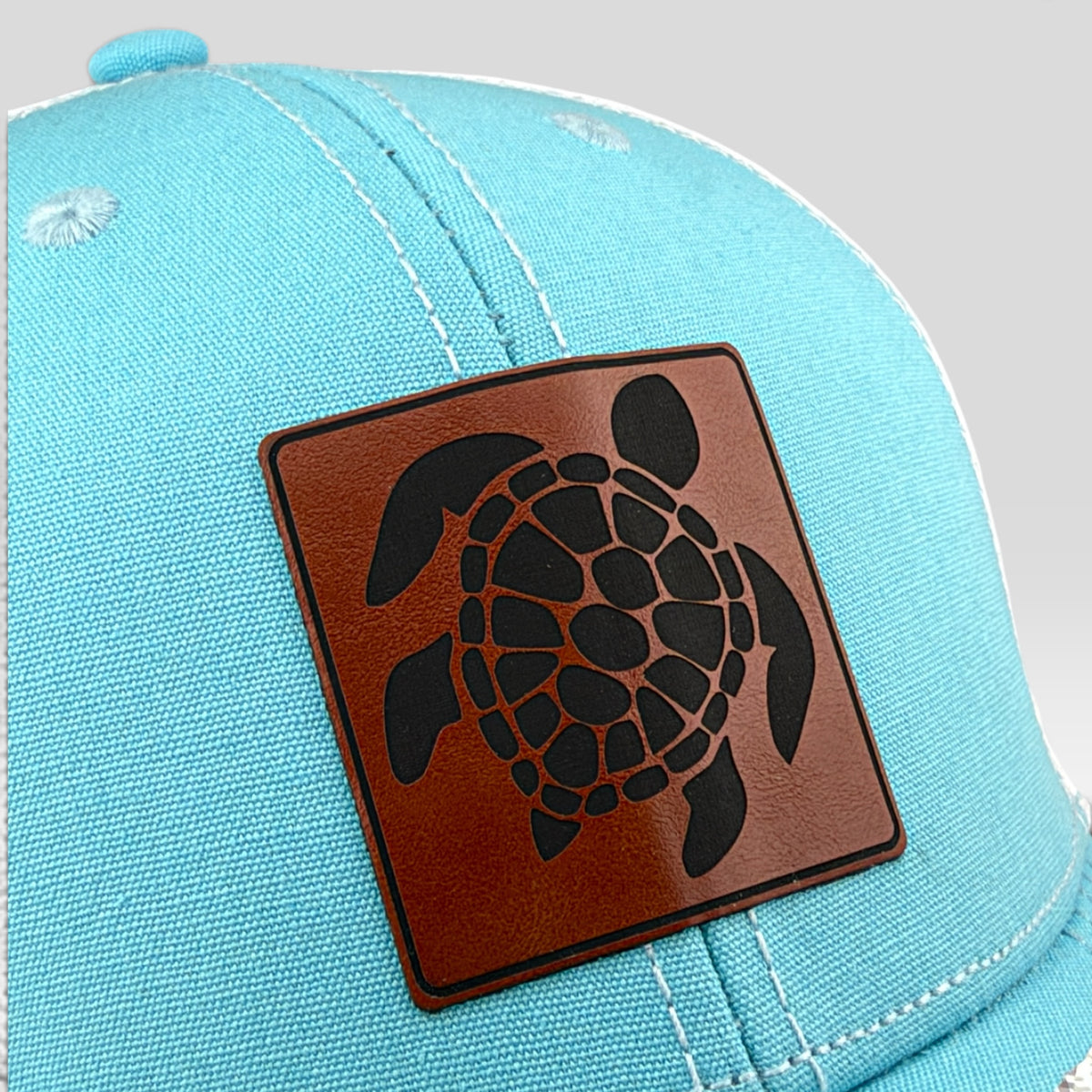 A close-up of a light blue baseball cap featuring a square brown leather patch with a black sea turtle design on the front.
