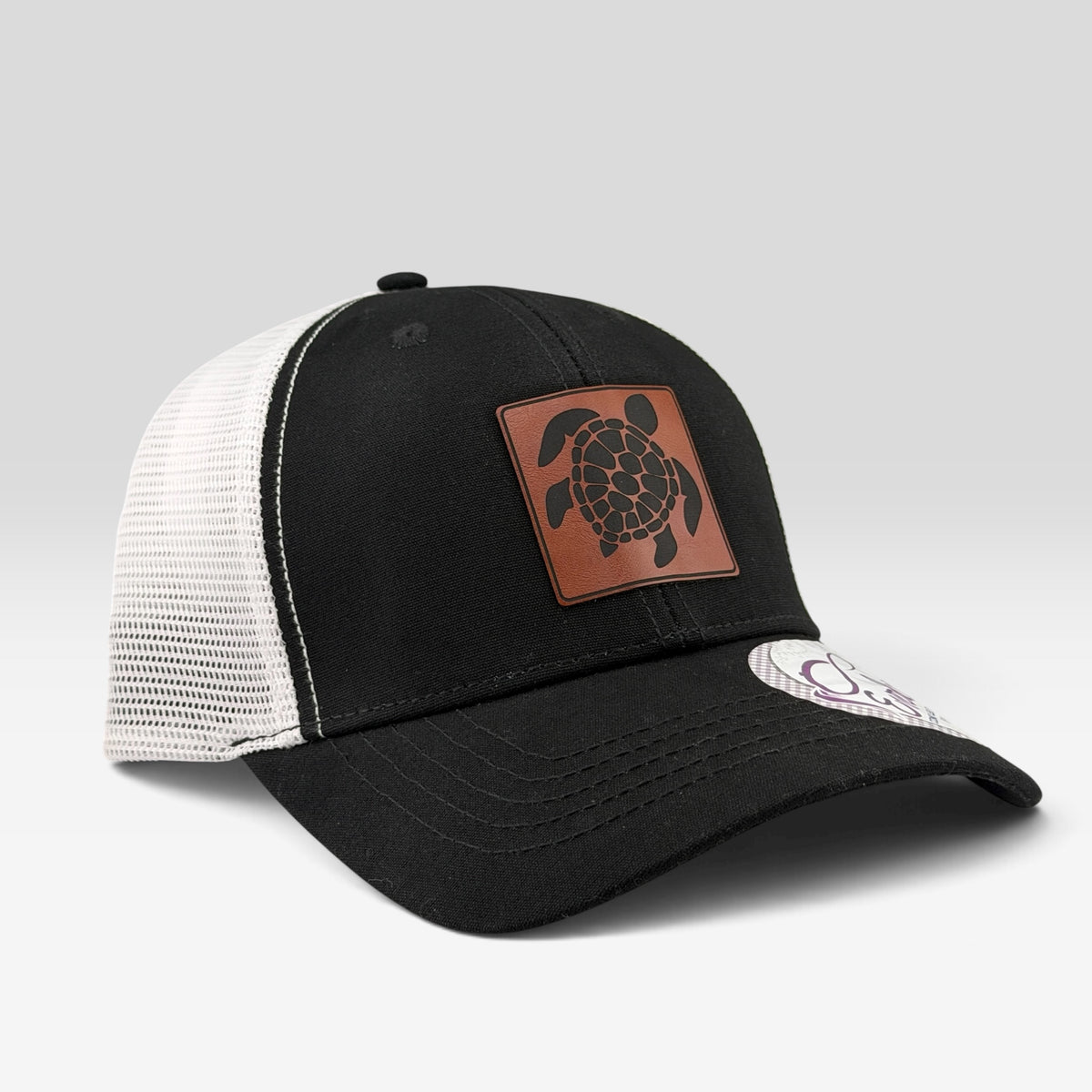 A black and white trucker hat featuring a brown leather patch with a black sea turtle design on the front. The hat has a black front panel, black brim, and white mesh back panels, photographed on a light gray background.