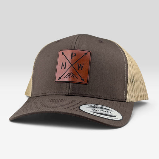 A brown and beige trucker hat with a brown leather patch on the front displaying 'P N W' with mountain and arrow details. A white circular sticker is visible on the brim.