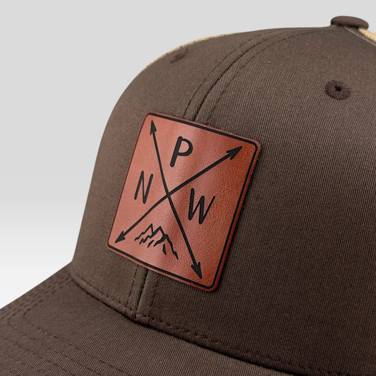 A close-up of a dark brown baseball cap featuring a square brown leather patch on the front. The patch displays a compass design with the letters 'P', 'N', and 'W' and arrows, with a small mountain graphic at the bottom.