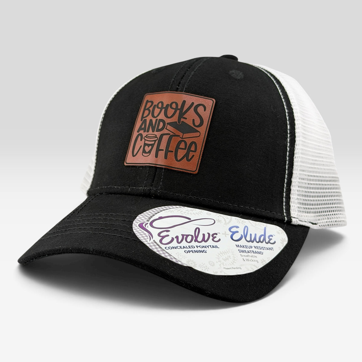 A black and white trucker hat with a brown leather patch reading 'Books and Coffee' on the front. The brim has a white sticker that reads 'Evolve Elude'.