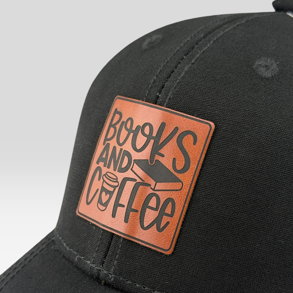 Close-up of a black baseball cap featuring a rectangular brown patch on the front with the words 'Books and Coffee' in black lettering and a small book-and-coffee graphic.