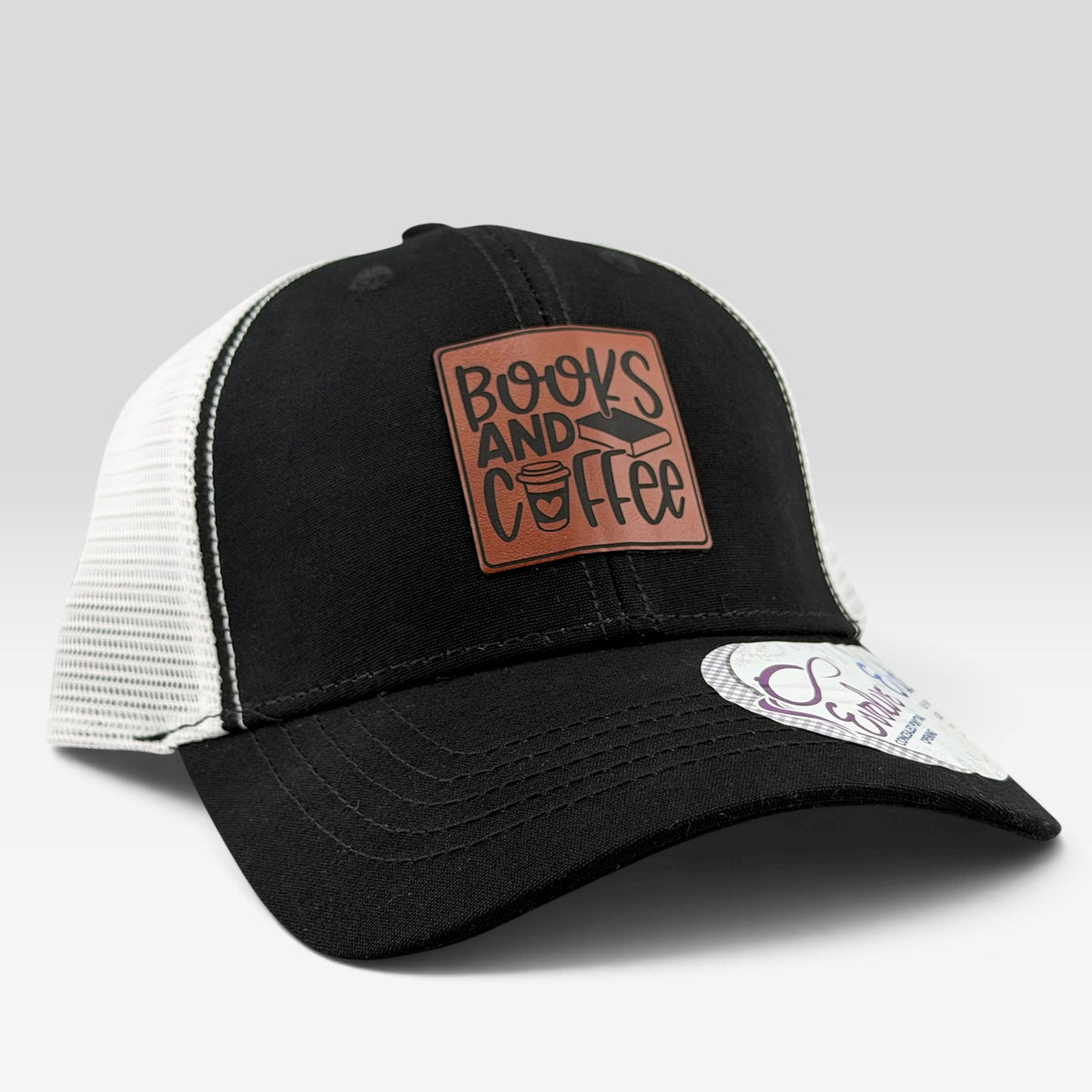A black and white trucker hat with a brown leather patch on the front that reads 'BOOKS AND COFFEE' in stylized lettering. The hat has a black front panel and brim, white mesh back panels, and a curved brim with a visible sticker on the side.