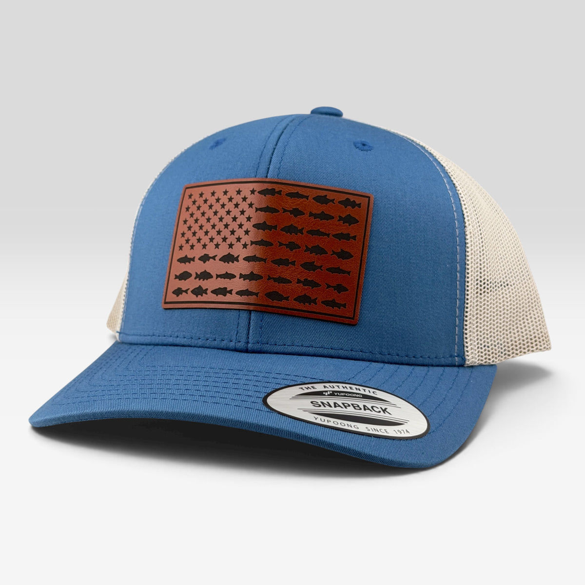 A blue and beige trucker hat featuring a brown leather patch on the front with a fish-patterned American flag design. The Snapback brand sticker is visible on the brim, set against a clean light gray background.