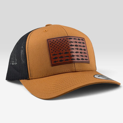 A tan and black trucker hat featuring a brown leather patch on the front with a black fish-patterned American flag design, photographed on a light gray background.