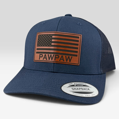 A navy blue trucker cap with a brown leather American flag patch on the front that reads 'PAWPAW'. The cap features a navy blue fabric front and brim, and a dark mesh back.