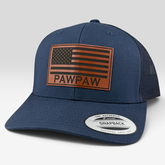 A navy blue trucker cap with a brown leather American flag patch on the front that reads 'PAWPAW'. The cap features a navy blue fabric front and brim, and a dark mesh back.