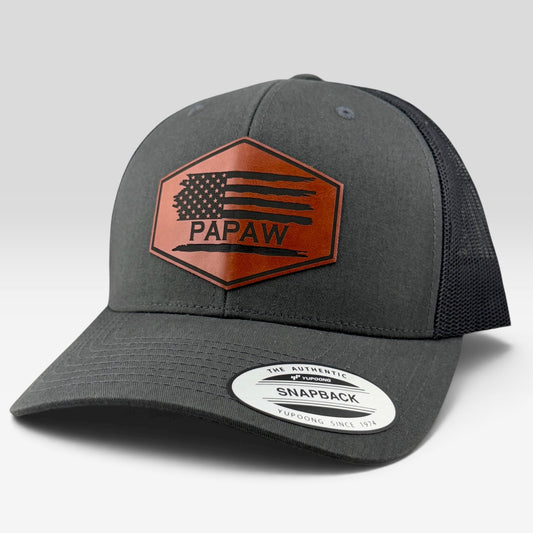 A dark gray and black trucker-style cap with a brown hexagonal patch on the front featuring a distressed American flag design and the word 'PAPAW'. A white Snapback sticker is visible on the brim, set against a light gray studio background.